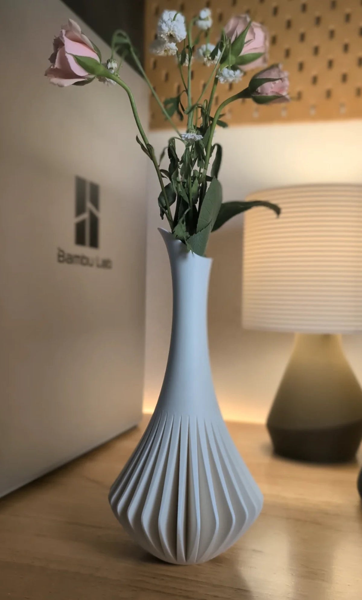 A white ribbed ceramic vase with a flared base, holding pink and white flowers, placed on a wooden surface in a softly lit indoor setting.